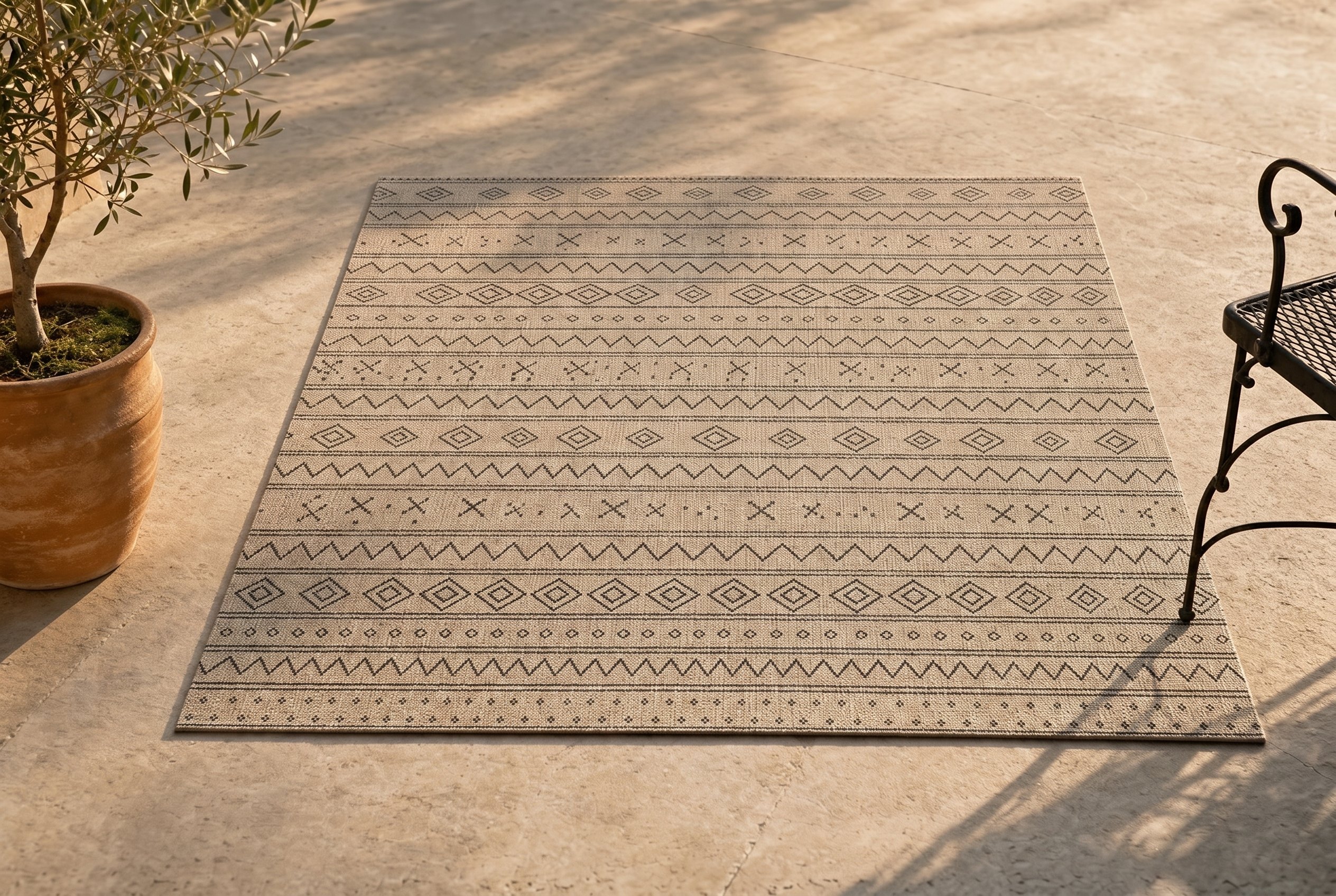 Boho outdoor patio rug in natural beige with Berber-style tribal stripe pattern of diamonds, zigzags, and X-motifs on a sunlit travertine stone patio, anchored by a terracotta-potted olive tree on the left and a black wrought-iron café chair on the right — boho-under-$80 outdoor rug lineup