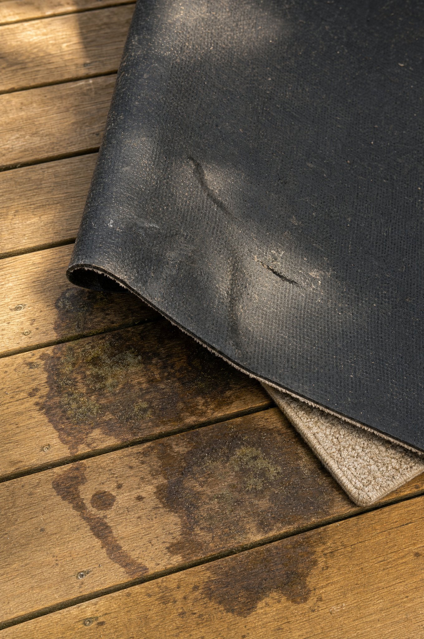Corner of a rubber-backed outdoor rug peeled back off a honey-toned wood deck, revealing dark water stains and discoloration in the wood grain underneath — the exact deck damage the 5-point check is designed to prevent