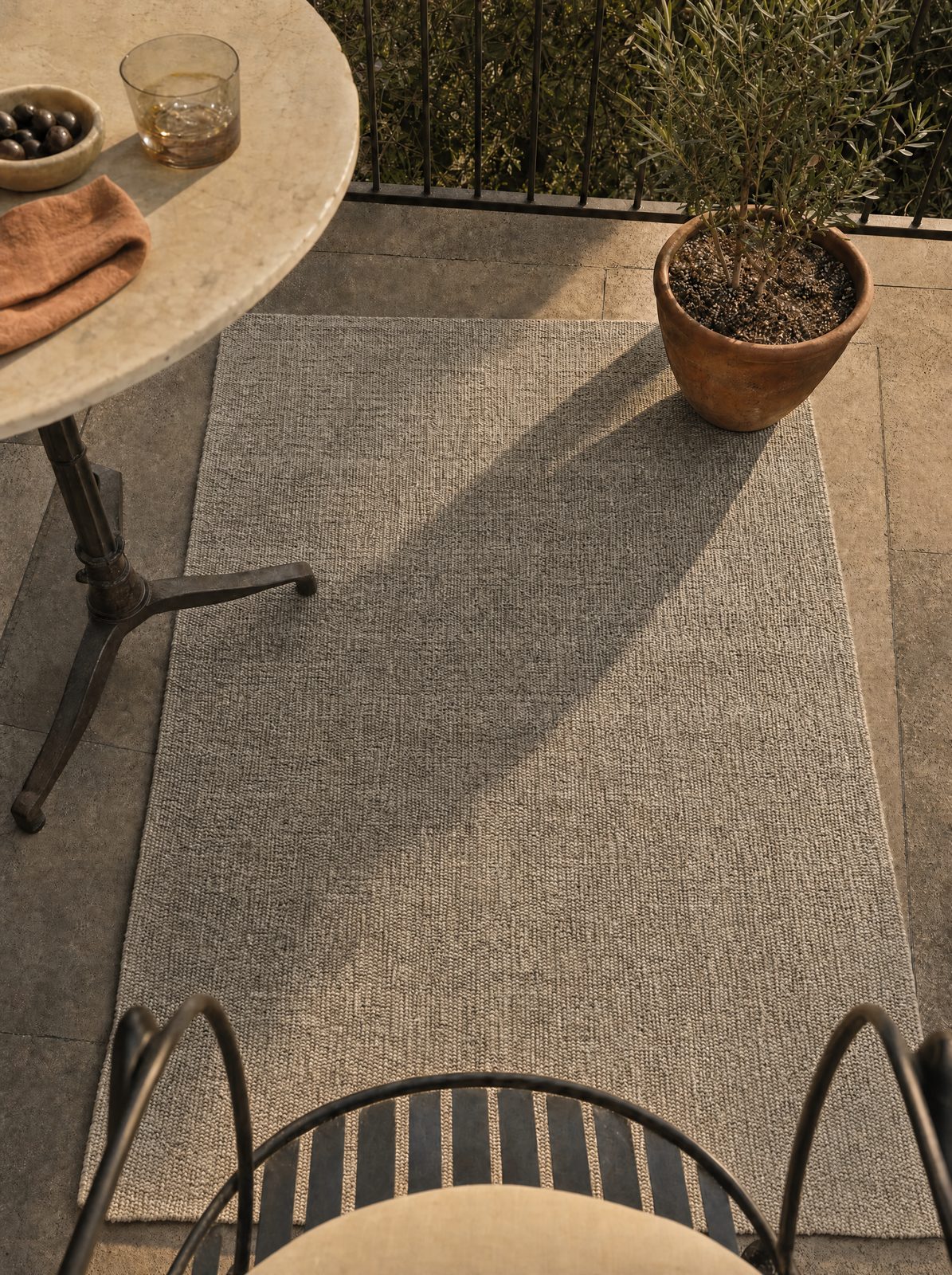 Unique Loom heathered beige-ivory flatweave solid outdoor rug at 4' 1" x 6' 1" on a stone balcony with marble bistro table, olives in a small ceramic bowl, terracotta-potted olive tree, and iron bistro chair